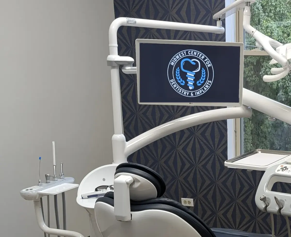 Treatment room at Midwest Center for Dentistry and Implants, with a patient chair, an overhead monitor showing the practice seal, and a window onto trees